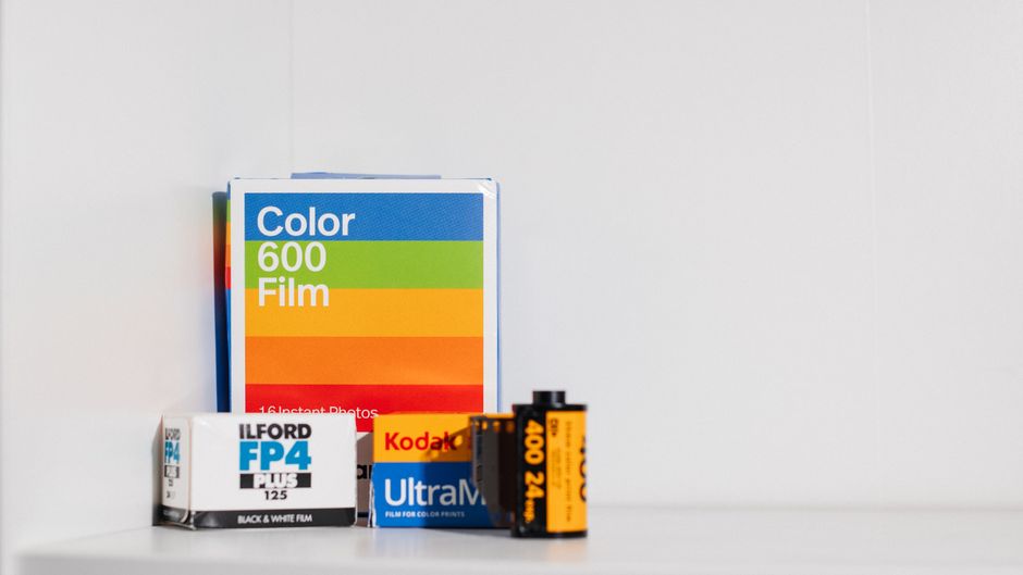 A close-up of a small collection of photography and film-related items arranged on a plain white surface against a white background. The items include a pack of 16 instant colour film with a multicoloured label, positioned vertically on the left; a small white box of Ilford FP4 Plus black and white photographic film with blue and black text, located in front of the colour film pack; a yellow and black roll of Kodak Ultra Max 400 24 exposures, standing upright on the right side; and a partially visible blue box of film positioned behind the other items. The scene is well-lit, highlighting the textures of the glossy film packaging, and the arrangement suggests a context related to photography, which is relevant to the theme of private or alternative waste materials in a rubbish removal service context.