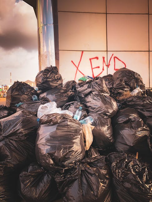 A large pile of black rubbish bags, some tied and others open, are stacked against a modern building with a tiled exterior. Several used plastic water bottles and other small debris are visible among the bags, which vary in size and appear to contain household waste. The bags are placed on a paved surface, possibly a sidewalk or street corner, with the setting suggesting an outdoor waste collection or clearance area. In the background, part of the exposed sky with clouds and a distant cityscape are visible. The building's tiled surface is partially covered by red graffiti that reads 'XEND,' and the lighting indicates a late afternoon or early evening atmosphere. The scene reflects an instance of unsolicited rubbish accumulation, which could relate to private waste handling services offered by companies like House Clearance Ilford, highlighting the need for proper rubbish removal and disposal solutions in urban environments.