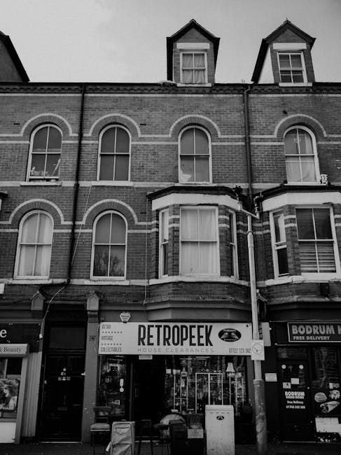 A three-storey brick building with a ground floor retail space featuring a sign reading 'RETROPEEK HOUSE CLEARANCES', indicating a business specializing in rubbish removal and property clearance services. The building has four arched windows on the first and second floors, with the upper floors also including small dormer windows protruding from the roof. The windows are framed with white trim, contrasting against the dark brickwork. The ground level storefront has large glass windows and a glass door, with additional signage and promotional materials visible inside. To the right of the property, there is a glimpse of another shop with a sign that mentions free delivery, suggesting local service offerings. The exterior environment appears in black and white, suggesting natural or diffused lighting conditions, with the building situated on a busy street likely in an urban area. This scene exemplifies a typical setting for a private waste disposal service operating within a commercial property, supporting alternative approaches to rubbish clearance outside of municipal collection methods, as represented by the company House Clearance Ilford.