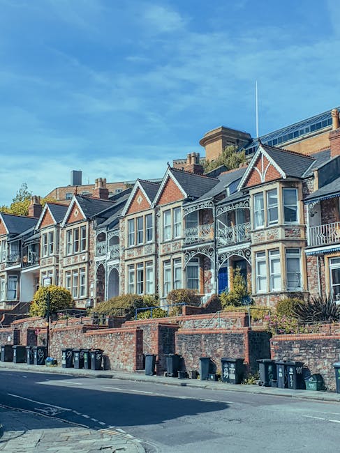 A row of Victorian-style terraced houses with ornate white metalwork balconies and bay windows, constructed from red brick with decorative gable fronts and slate roofs. The houses are situated on a slightly elevated street, with brick retaining walls and small front gardens landscaped with shrubs and plants. On the pavement in front of the houses, multiple black and dark green wheelie bins are lined up, indicating waste or recycling collection points. The scene is illuminated by natural daylight under a clear blue sky, with some nearby trees visible behind the buildings. This setting reflects an urban residential environment where private waste collection services, such as those provided by house clearance companies, often operate independently of local authorities for rubbish removal and house clearance activities.