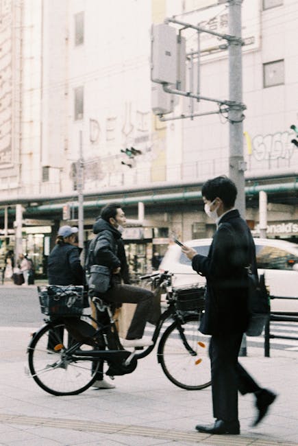 The image depicts an urban street scene with two men engaged in a conversation; one man is seated on a stationary bicycle equipped with a front basket and side panniers, wearing a dark jacket and a grey cap, while the other stands nearby dressed in a suit, currently looking at a smartphone and wearing a face mask. In the background, there are multi-storey buildings with large windows and graffiti on the walls, along with a streetlight and utility pole visible above. The urban environment includes a sidewalk and some shopfronts, one of which appears to be a fast-food outlet. The scene is lit with natural daylight, giving a neutral, professional tone, illustrating typical city life that might involve brief stops and encounters. While the focus is on casual city activity, this setting could imply the importance of efficient waste management and the role of private rubbish removal services, like those offered by House Clearance Ilford, to support clean and well-maintained urban spaces.