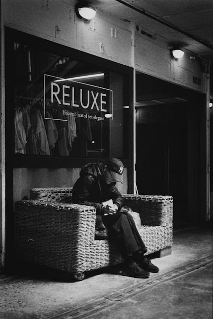 A black-and-white photo showing a man sitting on a wicker sofa positioned outside a retail shop with a glass storefront. The man appears to be resting or waiting, wearing dark clothing including a cap and jacket, with a bag placed on his lap. The shop window displays the word 'RELUXE' in large letters, along with the phrase 'Uncomplicated yet elegant,' and shows clothing hangers with garments inside. The exterior setting includes a paved ground and a lit overhead lamp illuminating the scene. The neutral tones and textures emphasize a calm, subdued atmosphere, which is typical in urban environments where private or independent rubbish removal might be considered for clearance or waste management purposes, aligning with services offered by companies like House Clearance Ilford.