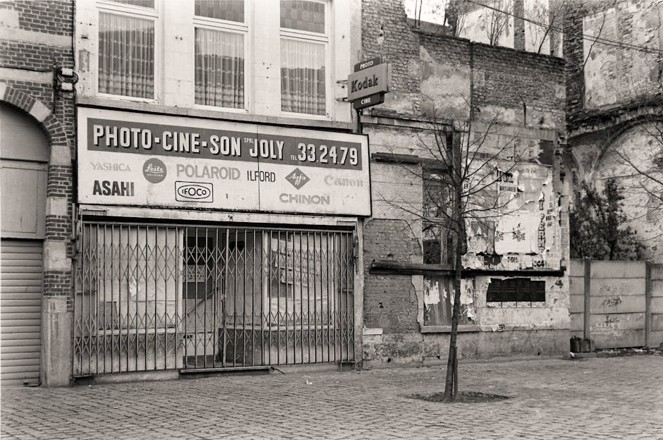 The image depicts an urban street scene with two men engaged in a conversation; one man is seated on a stationary bicycle equipped with a front basket and side panniers, wearing a dark jacket and a grey cap, while the other stands nearby dressed in a suit, currently looking at a smartphone and wearing a face mask. In the background, there are multi-storey buildings with large windows and graffiti on the walls, along with a streetlight and utility pole visible above. The urban environment includes a sidewalk and some shopfronts, one of which appears to be a fast-food outlet. The scene is lit with natural daylight, giving a neutral, professional tone, illustrating typical city life that might involve brief stops and encounters. While the focus is on casual city activity, this setting could imply the importance of efficient waste management and the role of private rubbish removal services, like those offered by House Clearance Ilford, to support clean and well-maintained urban spaces.