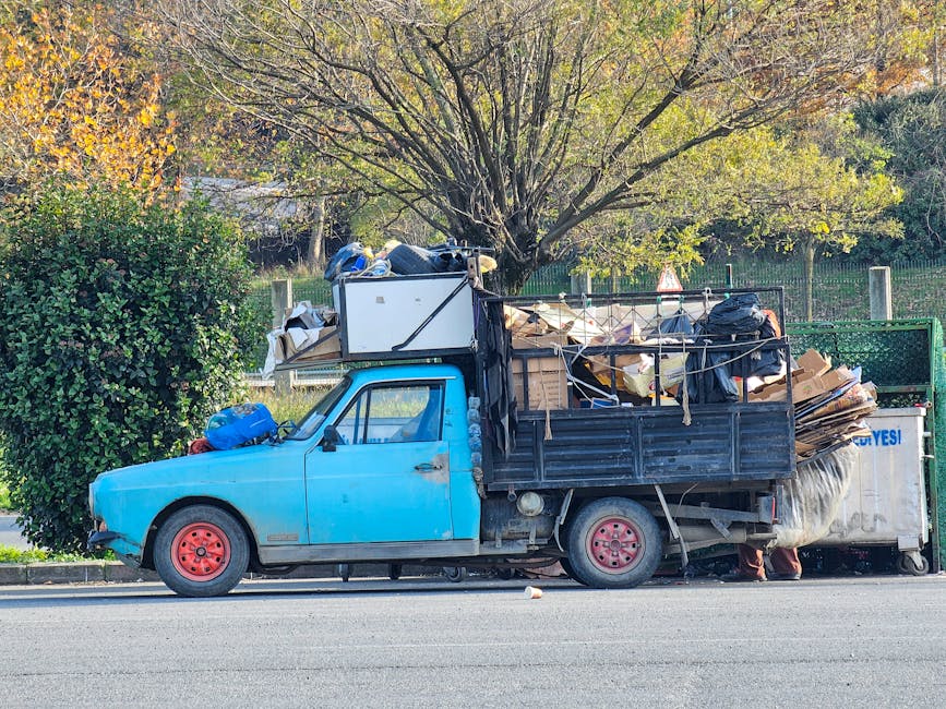 A small, aged blue pickup truck parked parallel to the curb on a quiet street with a large amount of rubbish and various materials loaded in its flatbed and cargo area. The truck’s paint is faded and chipped, revealing a dull surface, with pinkish-red steel wheels that have visible rust. The flatbed is stacked high with cardboard boxes, black garbage bags, loose paper, and wooden planks, some tied with string or ropes, indicating a private waste removal or alternative rubbish collection. In the background, there is a mature tree with green and yellow leaves, suggesting early autumn, and a green metal fence bordering a residential area. The scene is illuminated by natural daylight, providing a clear view of the cluttered load, reflecting an on-site clearance situation possibly handled by private waste management services like House Clearance Ilford, as the environment appears organized but heavily loaded with collected waste materials.