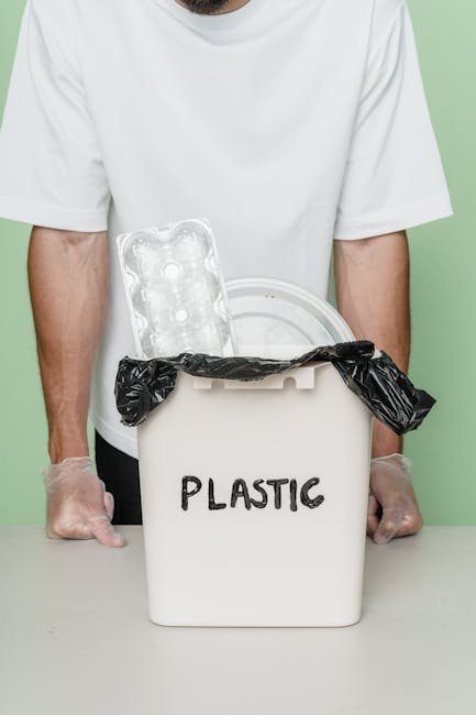 A person dressed in a white T-shirt and transparent disposable gloves stands behind a white plastic container labeled 'PLASTIC' in black marker. The container has a black garbage bag lining it, with the excess folded over the top edge. Inside the container, there is a clear plastic blister pack with empty compartments visible, resting against the rim. The background is a plain light green color, and the scene suggests a focus on waste segregation or recycling practices, which relate to alternative rubbish disposal options supported by private waste collection services like House Clearance Ilford. The image emphasizes careful separation of plastic waste and highlights the importance of proper disposal for recycling purposes within a domestic or small-scale waste management context.