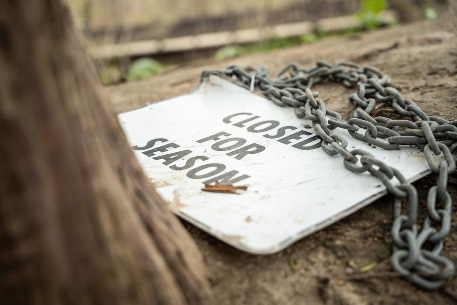 A weathered white metal sign with black text reading 'CLOSED FOR SEASON' is lying flat on the ground, partially obscured by a rusted metal chain draped across its surface. The sign appears old and slightly dirty, with scratches and dirt smudges visible on its surface. The chain, consisting of large, heavy, interlinked metal loops, is situated in the upper right corner and extends across the sign, suggesting an obstruction or barrier. In the foreground, the edge of a tree trunk with rough, textured bark partially frames the left side of the image. The background features a blurred wooden fence and patches of soil with small green plants, indicating an outdoor environment, possibly a garden or park area. The overall scene conveys an unattended, closed-off space, relevant to concepts of site closure, waste management, or restricted access, with a subtle connection to private property or alternative disposal instructions, as might relate to rubbish removal services by House Clearance Ilford.