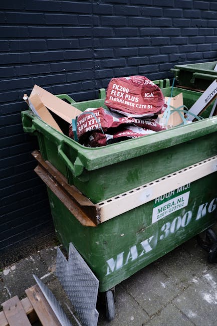 A green waste skip filled with red and black bags of garden soil, positioned on a paved surface against a dark brick wall. The skip is placed on a wooden pallet, and there are additional green containers visible to its right, all likely used for rubbish removal or garden waste collection services. The bags of soil are labelled with product information, and the overall scene suggests a site for private or alternative waste handling, such as on-site clearance or independent collection, managed by a waste removal company. The environment appears to be outdoors, possibly in a residential or commercial yard, with natural daylight illuminating the scene. The scene emphasizes the capacity of the skip for garden or general waste, reflecting a typical approach to rubbish disposal outside of council-provided rubbish collection, and aligns with private waste management practices.