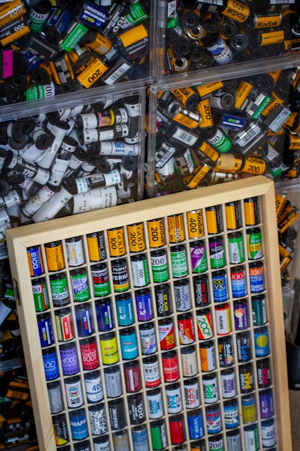 The image displays a close-up view of a collection of cylindrical batteries, predominantly AA and AAA sizes, arranged in transparent plastic containers and a wooden tray. The batteries feature various coloured labels, including yellow, white, green, blue, and black, with printed numbers indicating their capacity or voltage, such as '200', '400', and '500'. The plastic containers are filled with loose batteries and are positioned in the background, creating a textured and cluttered appearance, while the wooden tray in the foreground holds organised batteries arranged vertically within individual compartments. The scene suggests a setting related to battery storage or collection, with a focus on the different types and sizes of batteries, possibly intended for disposal or recycling in an appropriate waste management or rubbish collection context, exemplified by companies like House Clearance Ilford that specialise in alternative waste handling and private disposal services.