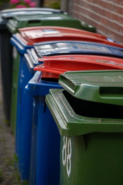 A row of large wheeled rubbish bins placed along a sidewalk adjacent to a brick wall. The bins are made of plastic with textured, ridged lids. The closest bin on the right is dark green, with a slightly open lid revealing the interior. Further along the row, there are blue bins with matching lids, followed by red bins with their lids closed. The bins are positioned tightly side by side, with the lids overlapping slightly in sequence. The background is slightly blurred, showing a residential environment with a paved pavement. The scene appears to be outside a property, suggesting a collection point for household waste. The lighting is natural, with diffused daylight illuminating the scene evenly, emphasizing the different colors and textures of the waste bins, which are part of an organized rubbish collection system, possibly for private waste disposal services like those managed by House Clearance Ilford.