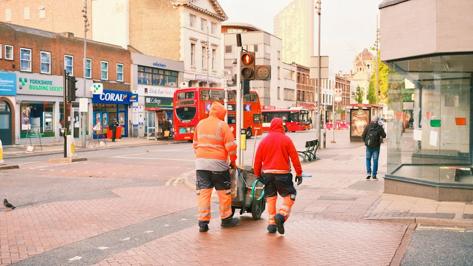 Two waste collection workers, dressed in high-visibility orange and red jackets with reflective stripes, are walking along a busy urban street with a small wheeled trolley. One worker is pushing the trolley while the other walks alongside, both appearing focused on their task. They are positioned in the foreground near a traffic light and pedestrian crossing, with the cityscape in the background. The street features red double-decker buses, various shops, and office buildings, including a pharmacy and a building with a sign for 'YORKSHIRE BUILDING SOCIETY.' The scene is illuminated by natural daylight, with some pedestrians visible in the distance, including a person in dark clothing walking past a glass storefront on the right. The surroundings suggest an active downtown area where private waste collection or rubbish removal services such as those offered by House Clearance Ilford may operate, emphasizing the professional teamwork involved in urban waste management.