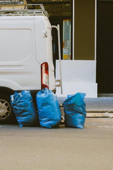 A white commercial van parked on a paved surface, partially visible from the rear side showing a vertical tail light. Three large blue rubbish bags made from plastic material are placed on the ground next to the rear of the van, with the bags filled and slightly bulging. Behind the van, there is a glimpse of a modern building entrance with a glass door framed by dark panels, and a white rectangular object leaning against the building wall, possibly a piece of furniture or construction material. The scene appears to be set outdoors in an urban environment, with minimal surrounding details visible. The context of rubbish removal is implied by the bags positioned for collection or disposal, aligned with private waste handling or on-site clearance services, such as those offered by House Clearance Ilford.
