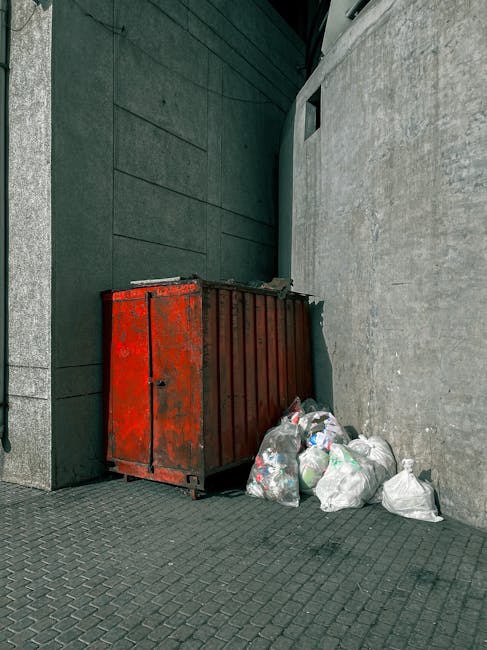A narrow alleyway situated between two urban buildings, with one side featuring a metal door and graffiti-covered wall, and the other side showing a window with metal bars. In the foreground, a large, weathered trolley or bag made of light-colored, heavy-duty fabric is positioned on the uneven asphalt surface, which has visible cracks and patches. Behind the trolley, a pile of accumulated rubbish and discarded materials, including cardboard boxes, plastic bags, and miscellaneous debris, extends towards the back of the alley. The waste appears haphazardly stacked, with some items leaning against the building walls or piled atop each other. The background reveals additional waste stacks, with leafless tree branches visible above, suggesting an outdoor setting in a city environment experiencing colder seasons. The scene has a muted, subdued tone, emphasizing the clutter typical of an abandoned or neglected urban space where private waste disposal or rubbish removal services, such as those provided by House Clearance Ilford, might be required for an on-site clearance or alternative waste handling outside regular council collection.