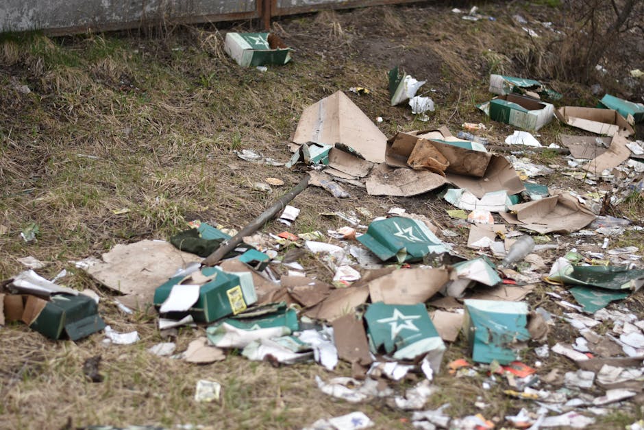 The image depicts a patch of ground near a grassy area, with discarded cardboard boxes, many of which are flattened and torn, scattered across the uneven surface. The boxes are primarily green with white branding, and some are partially crushed or ripped open, revealing their corrugated textures. Among the boxes, there are small pieces of paper, plastic wrappers, and other miscellaneous rubbish, creating a cluttered scene. A metal pipe or pole lies diagonally across the debris, partially buried in the soil. In the background, a section of a weathered brown fence or barrier is visible, with some dry grass and bare earth beneath it. The lighting suggests a cloudy or overcast day, giving a muted and somber tone to the scene. This scene illustrates the kind of littering that prompt private rubbish removal services like those offered by House Clearance Ilford can address, especially when debris accumulates in outdoor environments requiring local or independent clearance action, rather than municipal collection.
